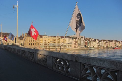 Rheinbrücke in Basel-Stadt mit Flaggen, Rhein und Gebäuden im Hintergrund.