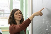 Musiklehrerin Imogen J. steht vor einer beschriebenen Wandtafel und zeigt auf Musiknoten.